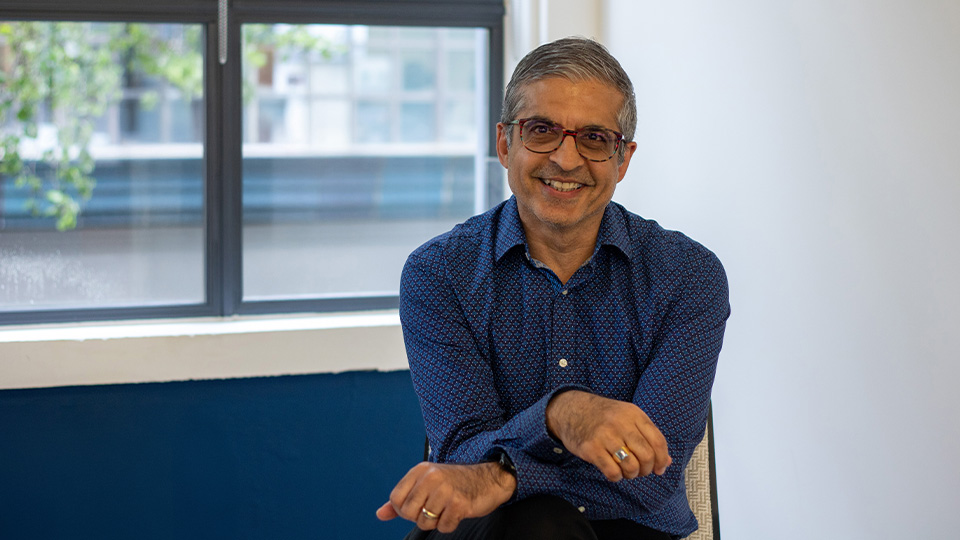 Deven leans forward in a chair, arms cross on his knees. He wears a navy shirt and brown glasses.
