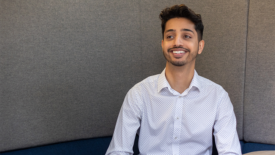 Indy Ghuman smiles off-camera. He wears a dotted white shirt. He is seated in an office space with grey and blue background