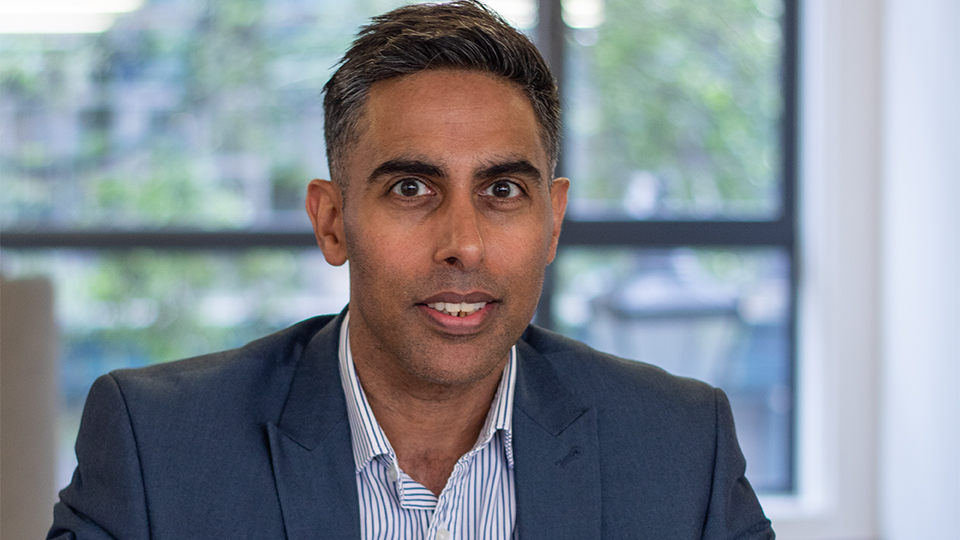 Kaljit is seated at a desk in the AOP office wearing a shirt and blazer