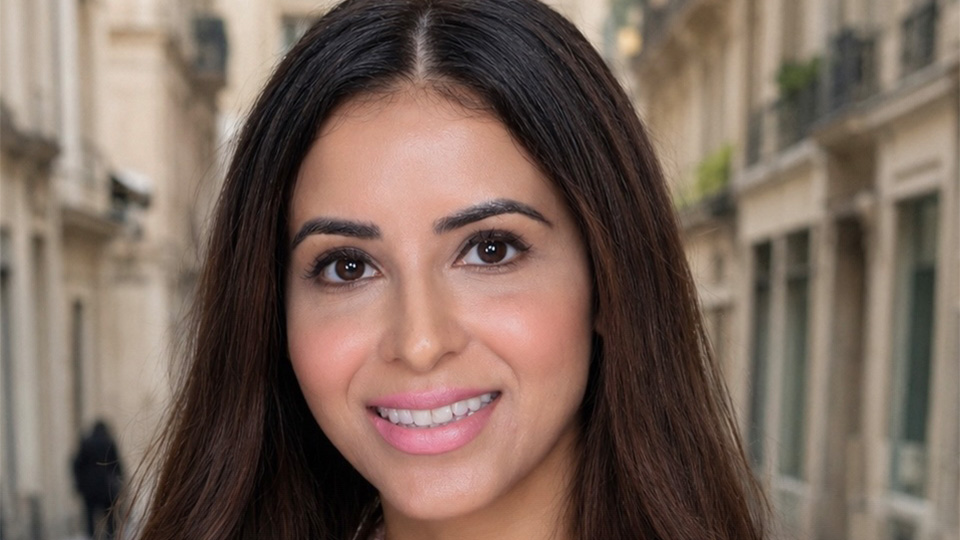 Kiran is photographed on a pale brick street, she wears a pink and pearl suit jacket and has long brown hair