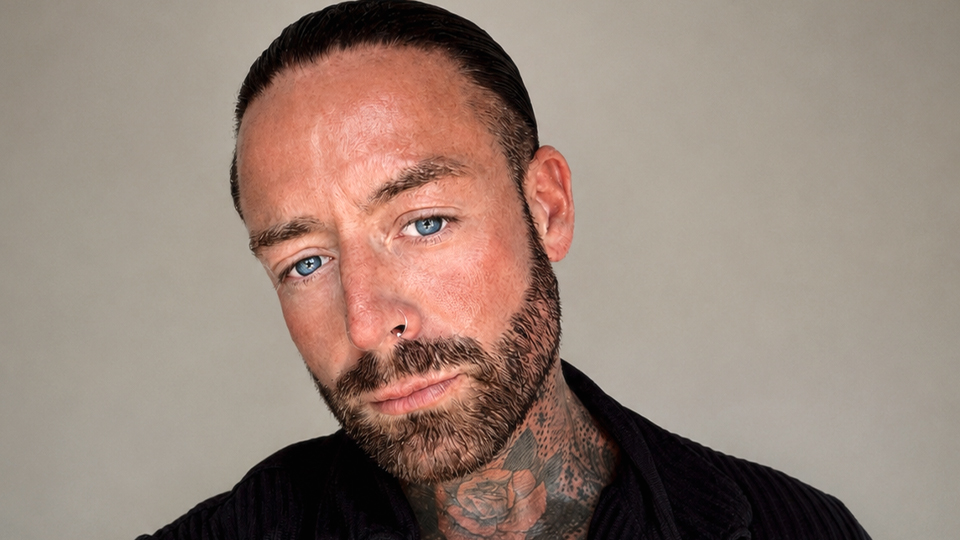 George poses for the camera with a neutral-serious expression. He has bright blue eyes, dark facial hair, and his dark hair is slicked back from his face. He wears a black textured shirt which shows his tattoos on his neck.
