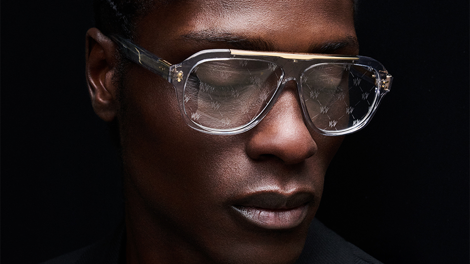 A model is photographed looking down in a black suit against a black background, throwing a highlight on their eyewear. The spectacles are an aviator style of clear acetate with a gold brow bar. Across the lenses are an impression of the William Morris London monogram