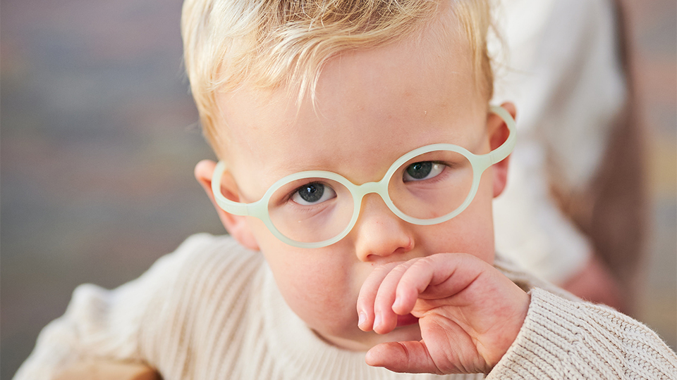 An infant boy looks shyly at the camera. He wears pale green glasses that sit snugly around his face