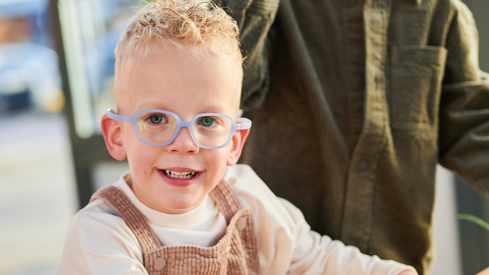 A pre-school age child wears soft square frames in a pale pastel blue. He wears brown corduroy dungarees over a long-sleeve t-shirt and is playing on a climbing frame
