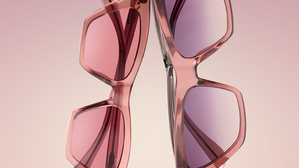 Two angular sunglasses in pink are balanced on their edges back-to-back on a pink display. One pair is smaller with pink lenses, while the larger pair is more pointed with a lavender lens