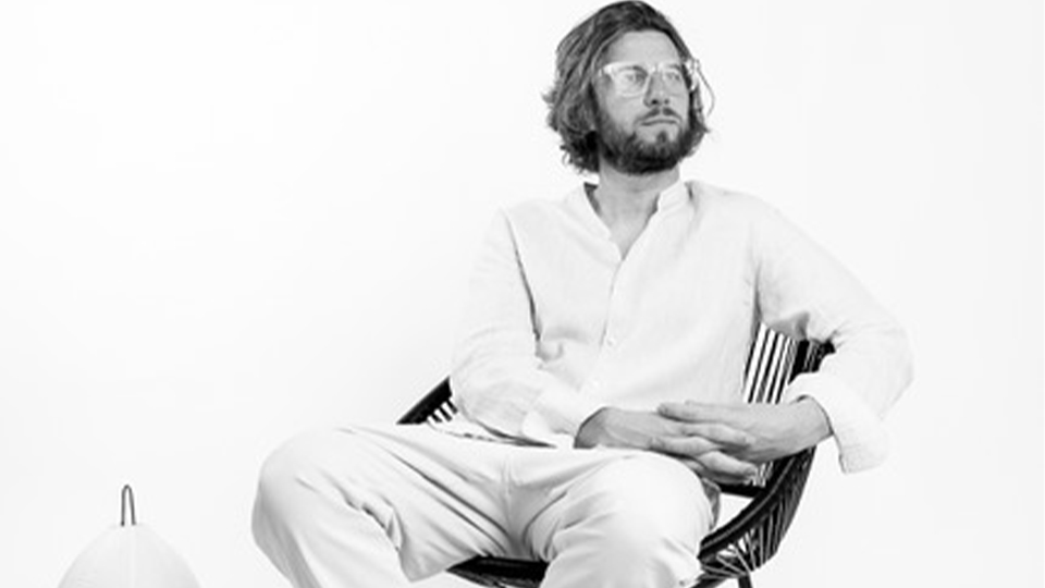 Marco is photographed in black and white. He wears a pale suit, sandals, and sits in a woven chair looking to the side. Beside him is a paper lamp and a Tree Spectacles book