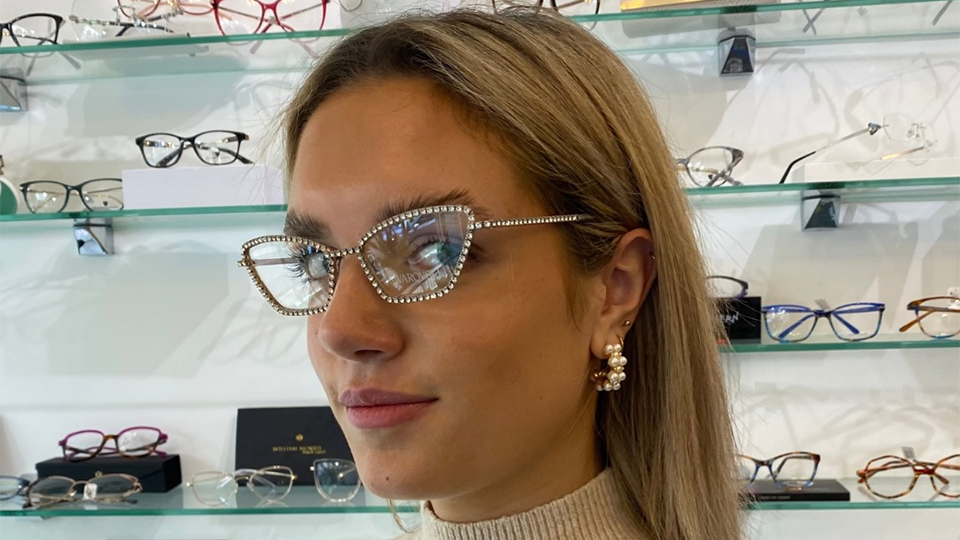 Meg poses in a practice with shelves of colourful eyewear behind her. She wears a sparkly cat’s eye style frame decorated with diamente