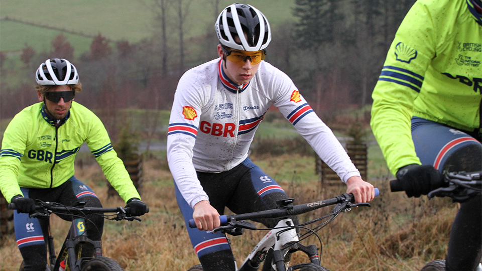 Three cyclists in sportswear with the GBR branding for the Great British Cycling Team in white and neon green, white helmets and navy shorts. The central figure wears sports frames with orange lenses. Behind him is another cyclist with black wraparound frames
