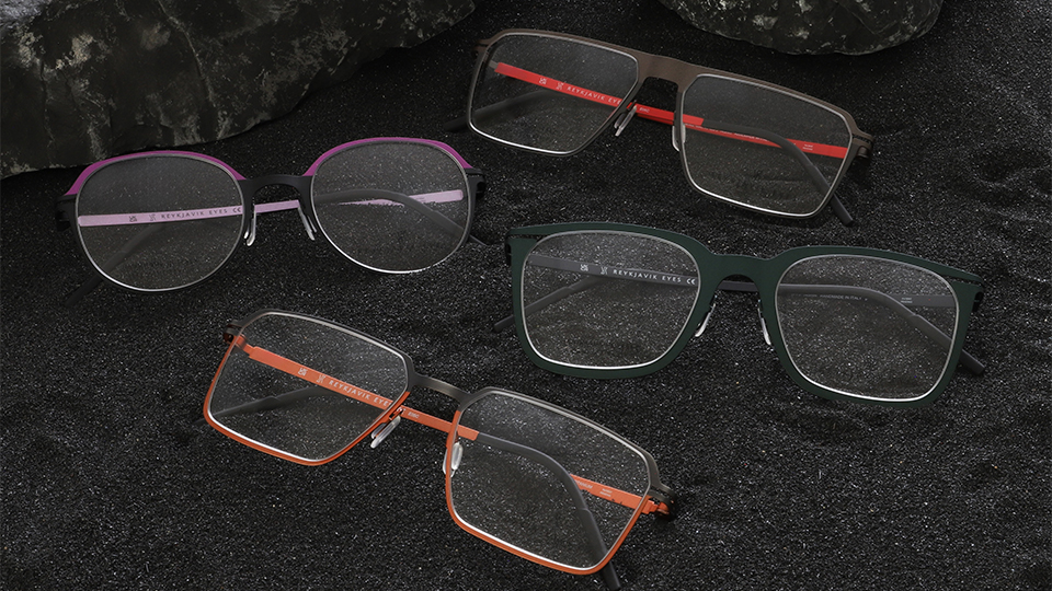 Four optical fames rest on a black sand and rocky beach. One of the frames has a purple browline and temples, another square frame has an orange lower half of the frame, a similar style has red temples, and a modern style is a deep green colour