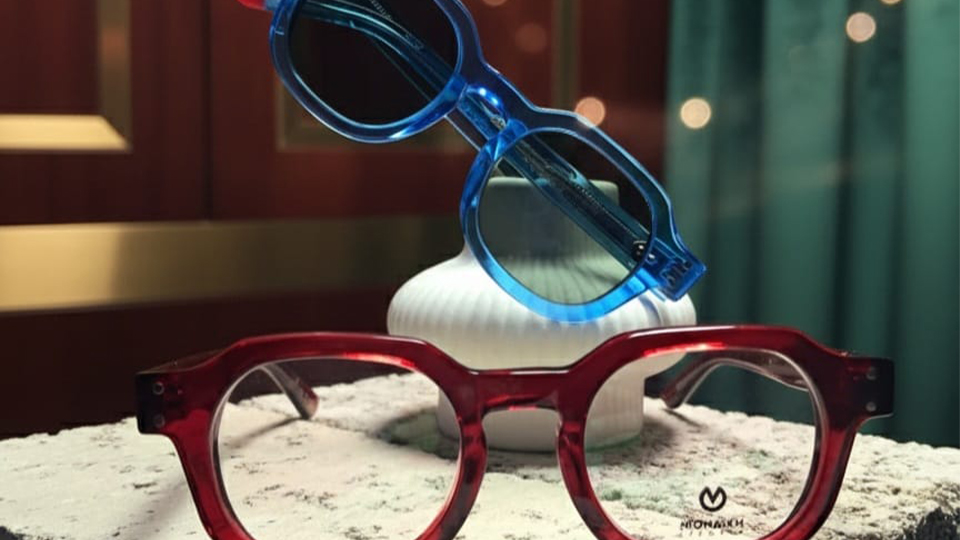A red frame is balanced on a rough concrete breeze block. Behind it, balanced in a white vase, is a blue frame. Both are in the pantos style with slightly flattened brows. The background is a fancy wood and green velvet backdrop