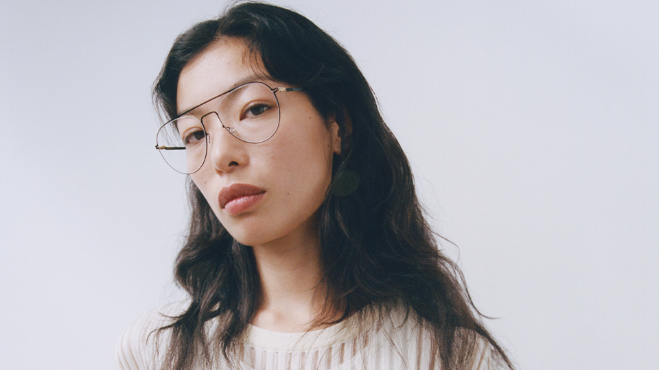 A woman wearing a cream knit one-piece with long sleeves models a pair of large thin metal frames in an oversized square style. The effect of the camera is soft