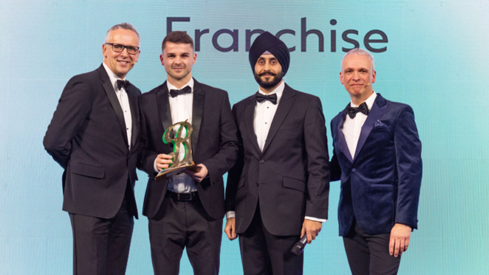 Four men in black tie stand on a stage at an awards ceremony, smiling for the camera. One man holds a trophy while another holds a microphone, with a large screen behind them displaying the word “Franchise”
