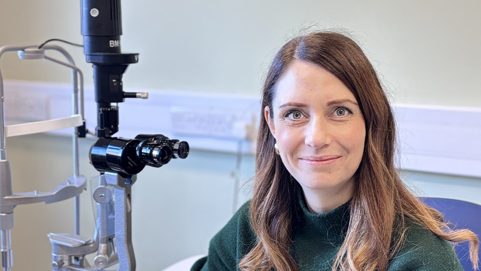 A woman with long brown hair sits beside a slit-lamp eye examination machine in a clinical room, smiling toward the camera. She is wearing a dark green sweater and is seated at a white desk with ophthalmic equipment