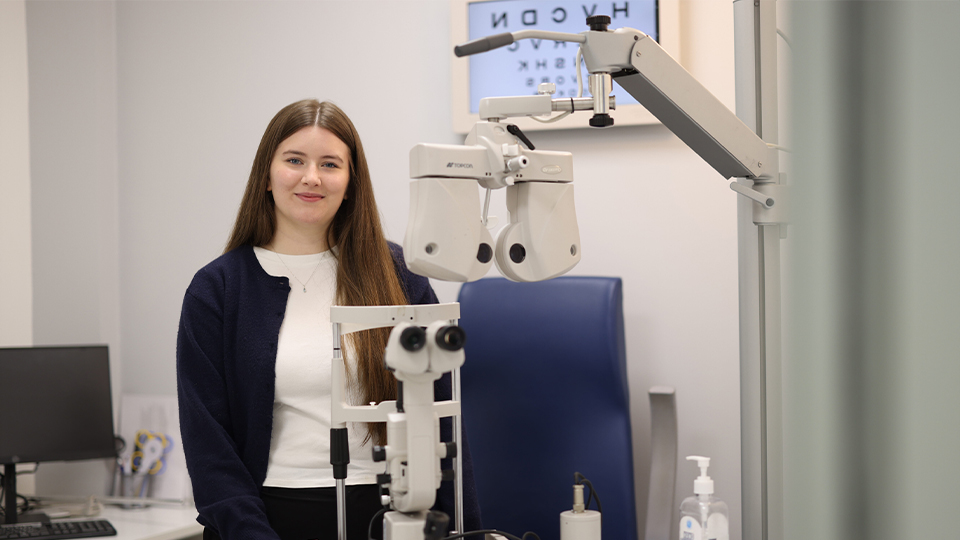 A woman with long brown hair stands in an eye examination room beside a slit lamp machine. She is smiling softly and wearing a white top with a dark cardigan. An eye chart is visible on the wall behind her, along with other optometry equipment, a computer workstation, and a bottle of hand sanitizer on the table