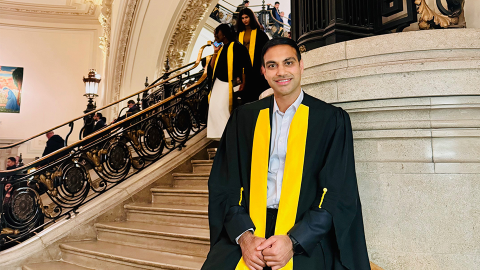 A man wearing a black graduation gown with a bright yellow stole smiles while standing on an ornate stone staircase inside a grand, historic building. Other graduates in similar attire descend the staircase behind him, framed by decorative arches, carved details, and wrought-iron railings
