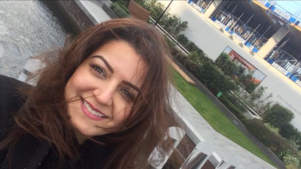 A smiling woman with long brown hair takes a selfie outdoors near a landscaped water feature with stone steps and greenery. Modern buildings with signage are visible in the background, and part of another person’s face appears at the edge of the frame