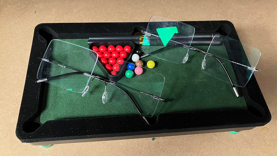 Two rimless frames with oversized square lenses are balanced on a mini snooker table