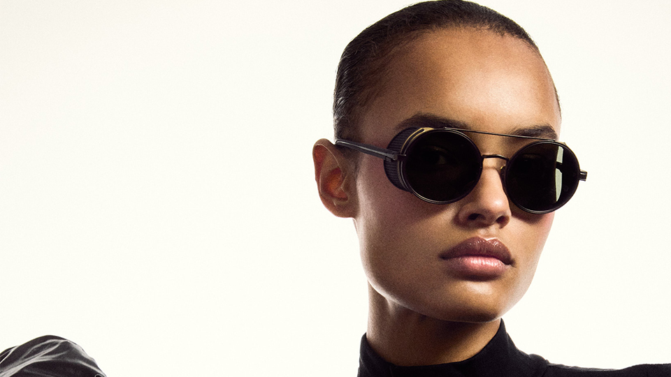 A model wearing black turtleneck and black puffer jacket wears large sunglasses with circular lenses and aviator-style metal detailing. The frames have gold side shields