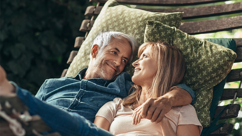 A middle aged man and woman sit together in a wooden hammock swing with plush cushions. They smile warmly at each other with arms around one another