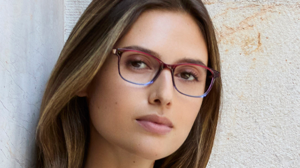 A female model wears slim rectangular glasses made of a plastic that graduates from red along the brow line to blue on the bottom of the frame. The model has brown hair past her shoulders and a v-neck navy linen top