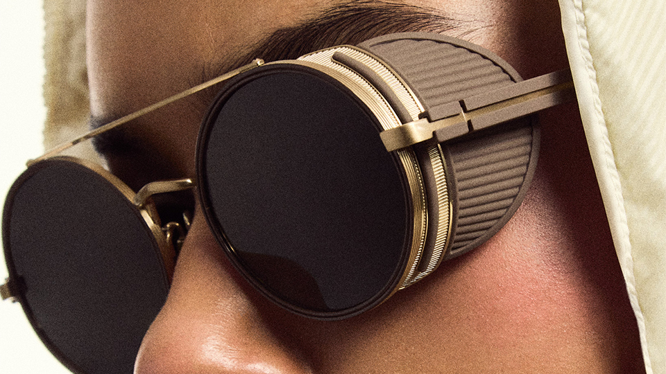 Close up of a model in a beige hoodie with large aviator style round sunglasses with side shields in gold and beige. The effect is modern and a little sci-fi
