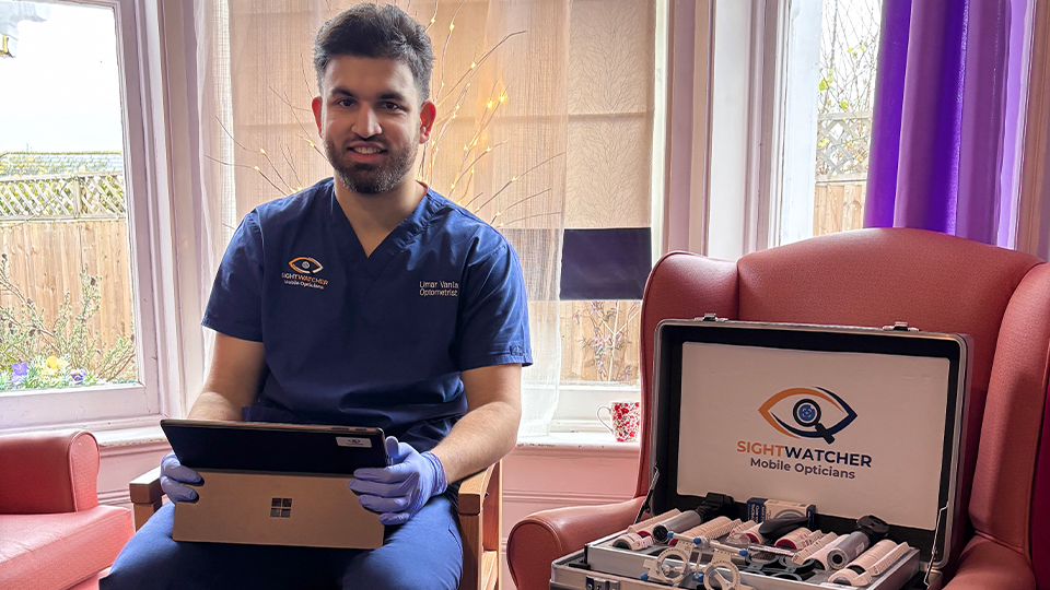 A man wearing blue medical scrubs and gloves sits on a wooden chair holding a tablet. He is indoors in a bright room with large windows behind him. Next to him, on an upholstered armchair, is an open silver case filled with optician tools and eyeglass frames, labelled “SightWatcher Mobile Opticians.”