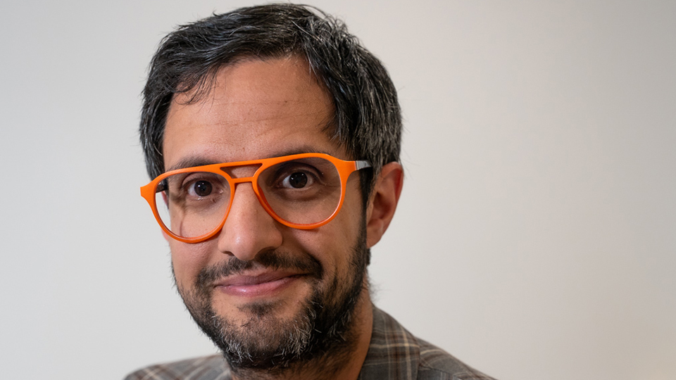 Close-up portrait of a man with short dark hair and light stubble, wearing round yellow glasses, a yellow T-shirt, and a blue-and-red plaid blazer. He looks directly at the camera with a slight smile, with a softly blurred indoor background
