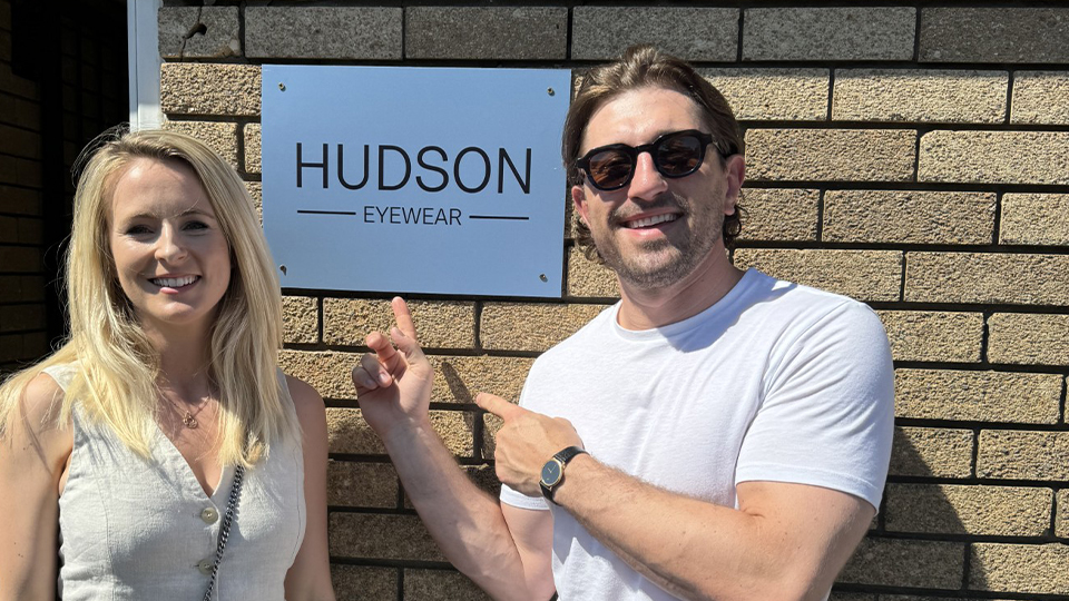 Two people pose by a sign for a warehouse stating Hudson Eyewear. The mood is celebratory