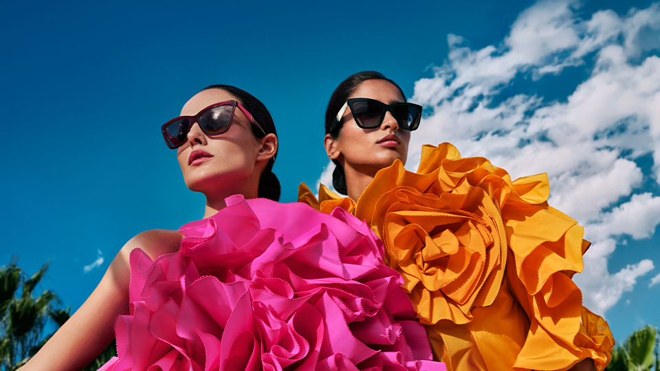 Two women photographed against a brilliant blue sunny sky in a classic country garden with stone vases and topiary. The women are dressed in yellow and lavender wrap dresses, hair slicked back into buns, and both wear oversized frames with thick sides in a marbled pink acetate