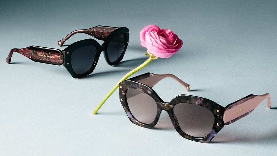 Two sunglasses are photographed with a bright pink flower. The frames are oversized, with thick chunky sides. The eyewear is made from a material combining many shades or pinks and purples. Large silver pins are a feature on either side of the frame