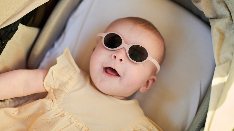 An infant in a summery yellow top and seated in a pushchair wears pale pink sunglasses that wrap around the head for a secure fit