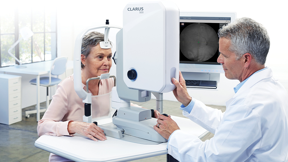 A patient, an older woman, is positioned against the chin and forehead rest of the Clarus 500, a piece of imaging equipment. On the other side, a clinician uses a joystick to prepare to take a scan