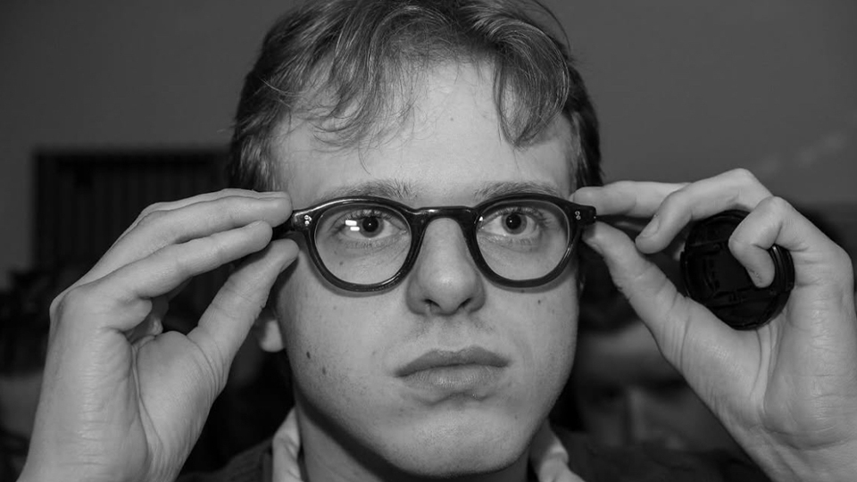 Jake is photographed in black and white adjusting a pair of dark spectacles on his face