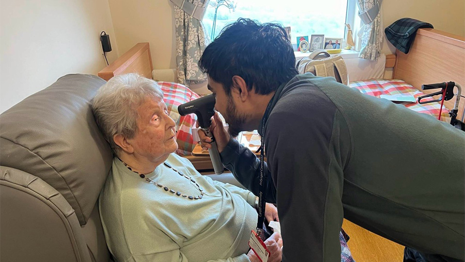 An optometrist leans in to examine an elderly woman’s eye using a handheld ophthalmoscope while she sits in a cushioned chair in a care home bedroom. A bed, walking aid, and personal items are visible in the background, suggesting a home visit eye examination