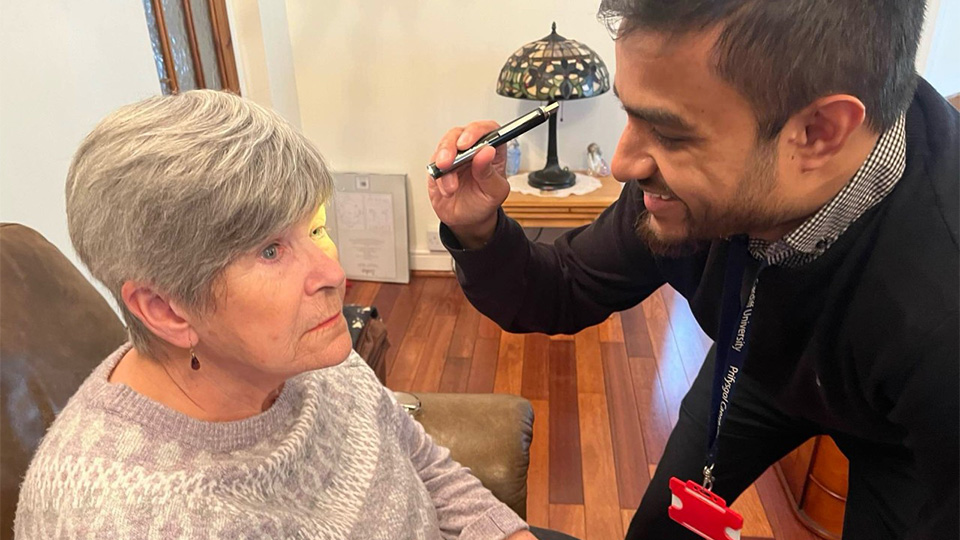 An optometrist shines a pen torch into an older woman’s eye while she sits in an armchair, looking upward during a home eye examination. The setting appears to be a living room, with wooden flooring, a side table, and a decorative lamp in the background