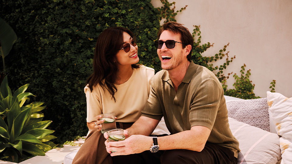 A young couple sit together in a summery terrace surrounded by plants and shurbs. Both wear neutrals in beige, browns, and khaki. Both wear sunglasses