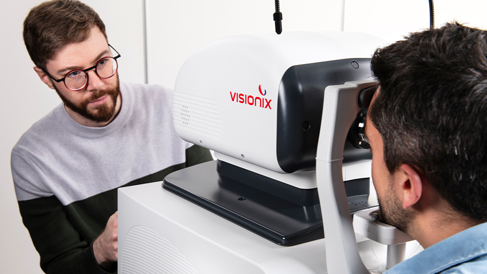 A patient leans forward to have an eye examination, their chin is on the headrest for the machine. On the other side, a clinician prepares to take the scan