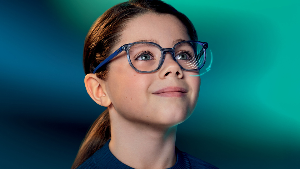 A young girl wearing spectacles is photographed, smiling hopefully upwards, against a blue-green background