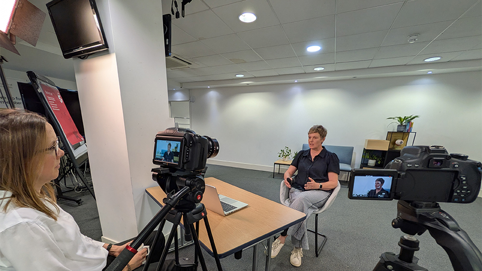Ceri Smith-Jaynes and Nicky Latham sitting in a room either side of a table with two cameras pointing at them
