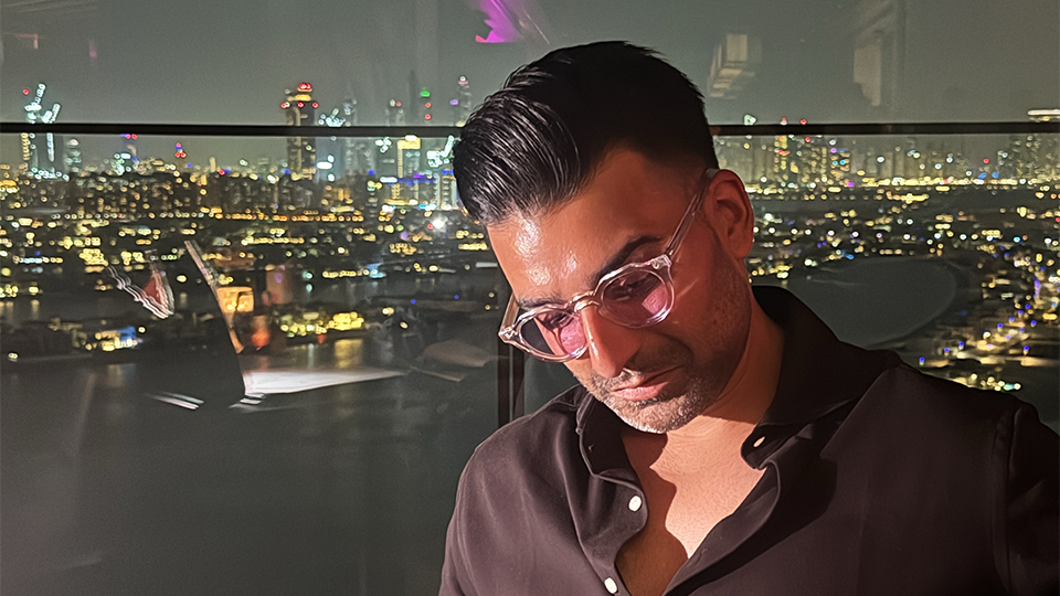 Vik Malhi is seated in a rooftop bar at night, the lights of a city glowing behind him. He wears a pair of translucent frames with pink tinted lenses