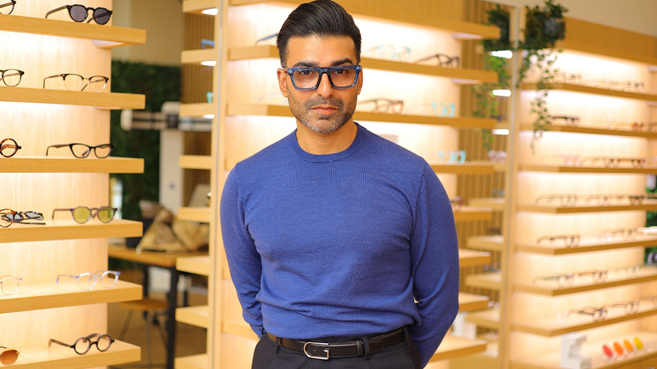 Vik stands in an optical practice with walls of warm wooden displays. He wears bold rectangle spectacles in a deep royal-blue, matching his jumper. He looks seriously at the camera