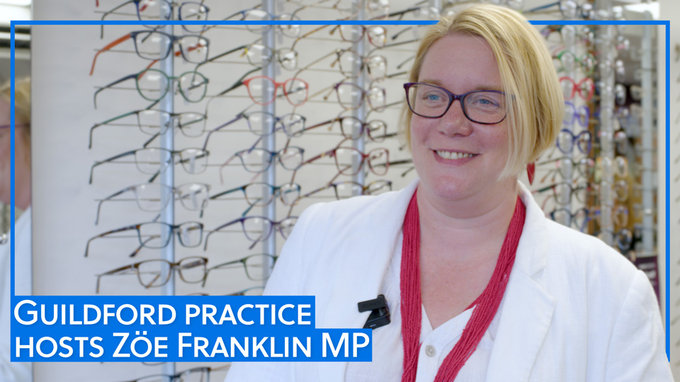Zoe Franklin, MP for Guildford, stands in an optometry practice. She wears a white blazer over a shirt and a bright red beaded necklace. Zoe wears glasses and has a blonde bob