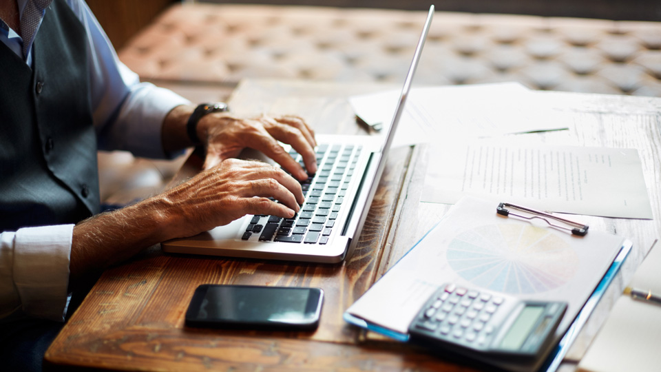 Elderly unrecognizable entrepreneur using a laptop. Mobile phone, a calculator, a pie chart and some papers and notes are on the table. 