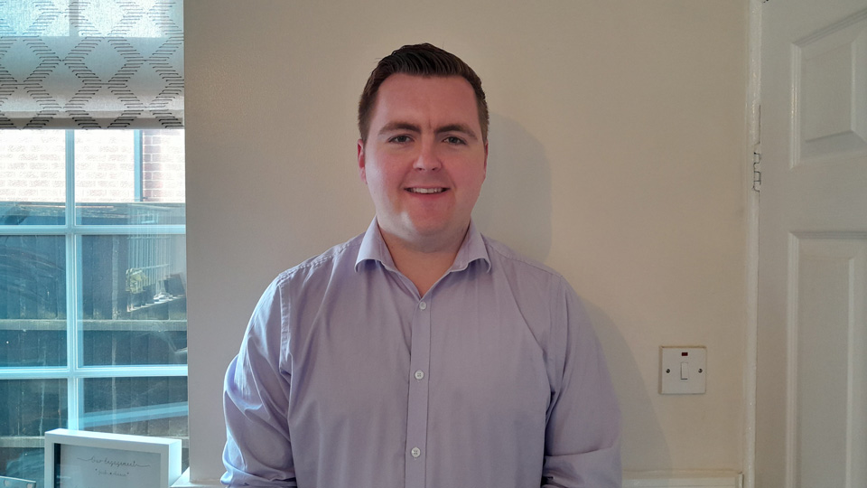 Jack stands smiling in front of a cream and green wall. He wears a light purple shirt, and his dark hair is in a short quiff