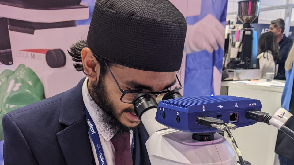 Male student, Habeeb Rahman, wearing a navy suit with a burgundy tie and wearing glasses. He is leaning in to look through the eye piece of an optical instrument. Around his collar is a 100% Optical lanyard 