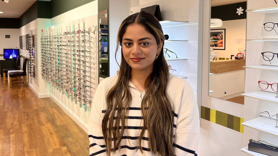 Aisha stands in an optometry practice, rows of spectacle displays behind her