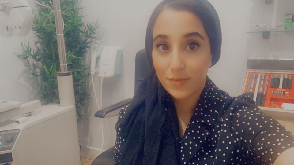 Aneesa is sitting in an optical practice room. She's wearing a black shirt with white polka dots and also is wearing a black head scarf 