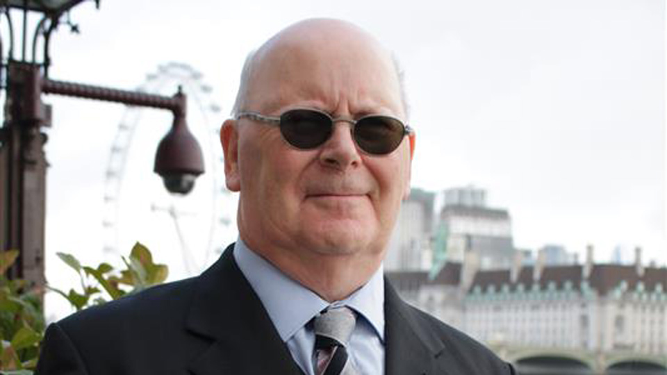 Elderly man with dark sunglasses smiling wearing black suit jacket, pale blue shirt and tie. In the background the London Eye wheel can be seen. 