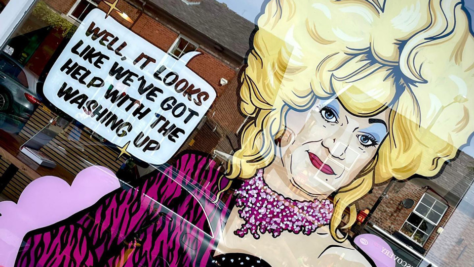A window display shows an illustration of the drag queen Lily Savage with blonde hair, a pink dress and blue eye make-up. A speech bubble next to Savage says, ‘Well it looks like we’ve got help with the washing up.’ 