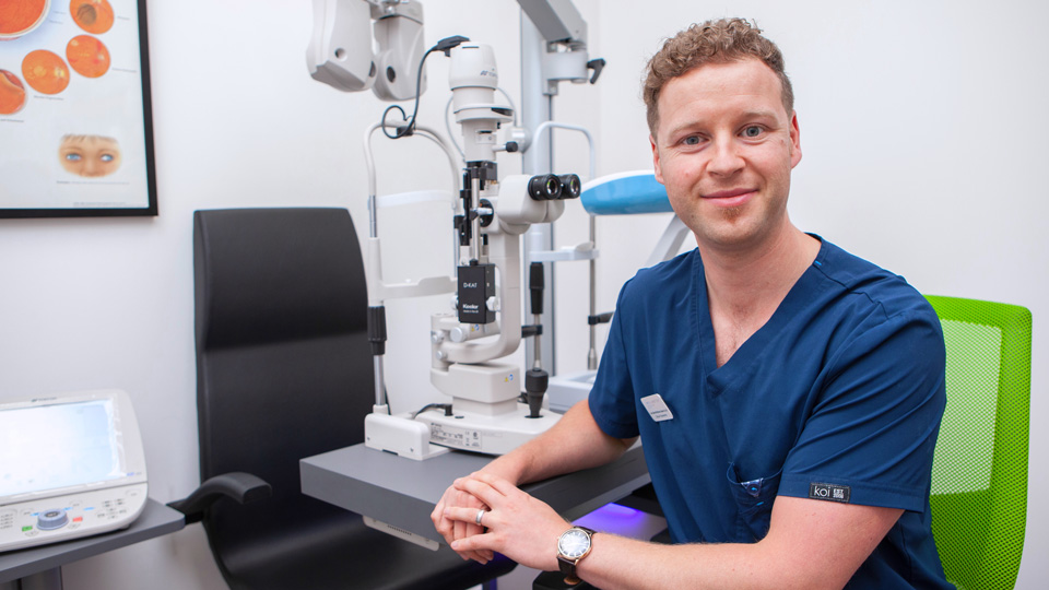 A man in dark blue t-shirt sitting in an optical practice smiling